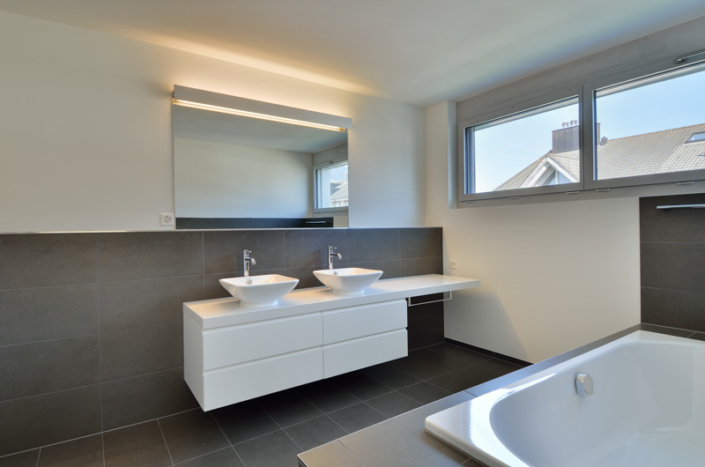 Salle de bain avec sol et moitié du mur sombres, doubles vasques avec meuble de rangement blanc en dessous, miroir à led au dessus