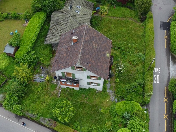Vue aérienne sur maison au toit mensardé, façade blanche et volets verts, entourée de verdure